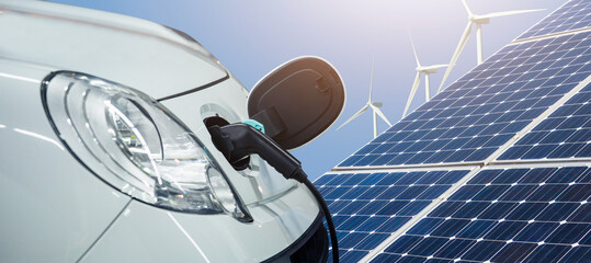 Close up of electric car with a connected charging cable on the background of solar panels and wind turbines - sources of clean renewable energy