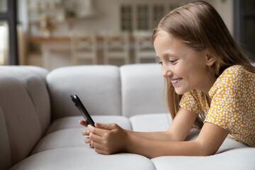 smiling little child lying on soft couch at living room look at phone screen use game app contact ma