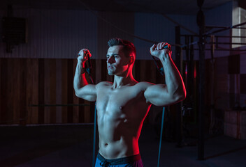 Wall Mural - Muscular powerful man with naked torso trains with expander (fitness rubber bands) in red blue neon light. Cross workout in modern gym