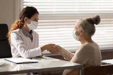 Wall Mural - Side view smiling female doctor wearing face mask and mature patient shaking hands at meeting in office, senior woman making health insurance deal, successful medical checkup results, healthcare