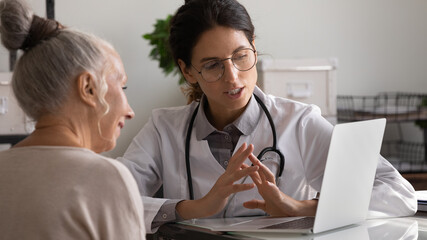 Wall Mural - Close up professional female doctor wearing uniform and glasses consulting mature patient, talking, using laptop, explaining prescription, discussing treatment, medical checkup result in hospital