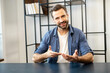 Young bearded hipster guy in casual clothes sitting at desk, looking at camera, moving hands, telling someone to introduce tell more about yourself or presenting the idea while on business video call