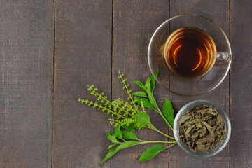 Tulsi or holy basil tea in transparent glass cup with fresh tulsi leaf and dried leaf in transparent glass bowl on wooden background, top view. Ayurvedic medicine in India.