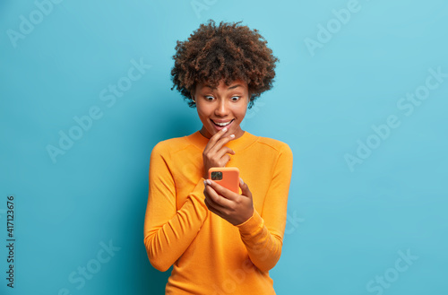 Happy surprised Afro American woman browses internet and networking in social media tests new application for smartphone wears casual jumper isolated over blue background. Mobile communication © Wayhome Studio