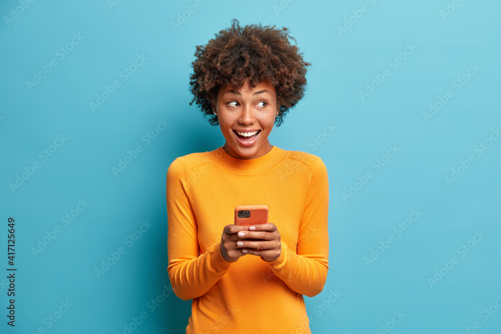 Positive curly haired ethnic woman uses mobile phone checks messages and reads news holds modern cellular in hands looks with curious happy expression on right isolated over blue background. - obrazy, fototapety, plakaty 
