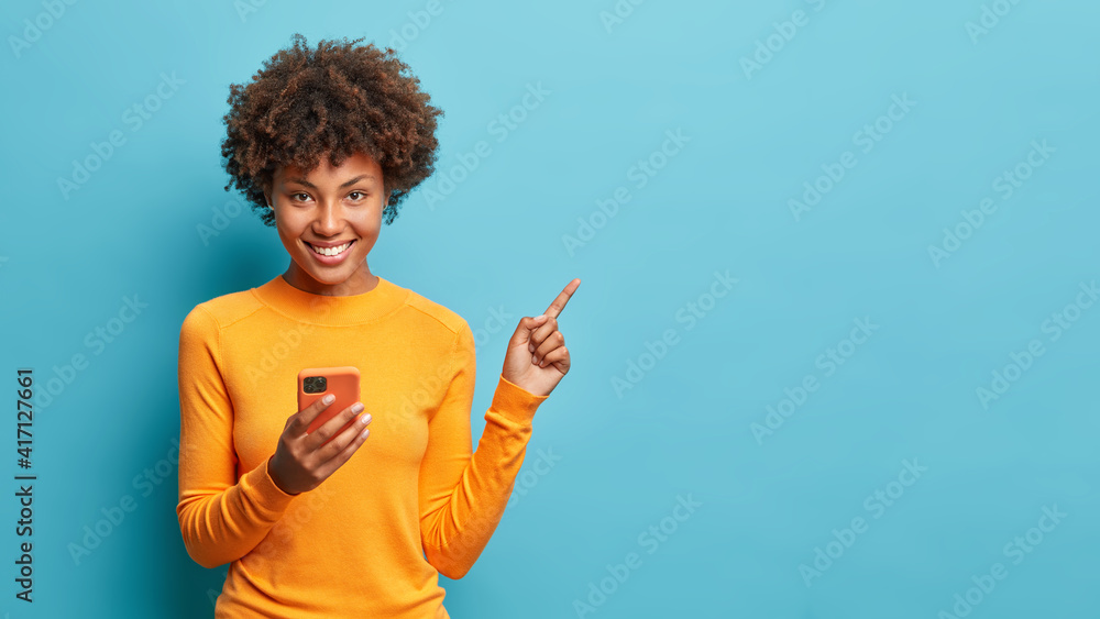 Horizontal shot of happy dark skinned Afro American woman enjoys mobile communication and modern technologies poses against blue background points away on free space for your advertising content - obrazy, fototapety, plakaty 