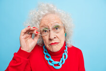 Portrait Of Beautiful Wrinkled Senior Woman Has Curly Grey Hair Bright Makeup Manicure Looks Attentively Through Transparent Glasses Wears Red Jumper And Necklace Isolated Over Blue Background.