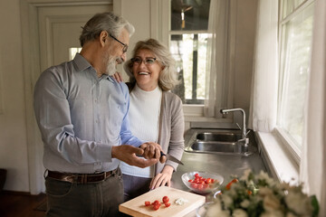 Happy middle aged couple of vegans cooking dinner together at home, making raw organic salad. Senior retired husband and wife cutting vegetables in kitchen, having fun talking and laughing