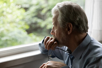 Pensive mature 70s aged man in deep thoughts holds glasses, looks out window. Thoughtful older person feels concern, depressed elder thinks of health problem and loneliness, suffers from memory loss