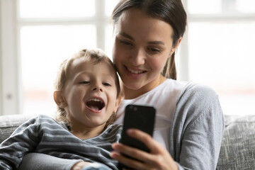 Wall Mural - Happy mum and excited cute son kid speaking and smiling at smartphone screen, resting on sofa at home. Mother hugging child, using mobile phone for video call, taking selfie, shopping online