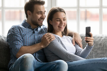 Wall Mural - Happy millennial husband and wife using mobile phone for video call, having funny talk. Young couple sitting on sofa, looking at cellphone screen and laughing, browsing online app for shopping