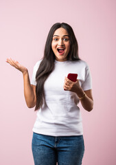 Smiling Indian young woman wears white T Shirt using smartphone or cellphone against pink background