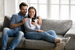 © fizkes - Happy millennial married couple with cellphone relaxing and hugging on couch, using online app on smartphone. Young man and woman with mobile phone making video call from home, shopping on internet