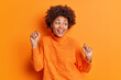 © WHstudio Leushin N - Optimistic dark skinned woman dances with raised arms moves carefree smiles broadly dressed in casual jumper isolated over vivid orange background celebrates achievement and victory enjoys nice day