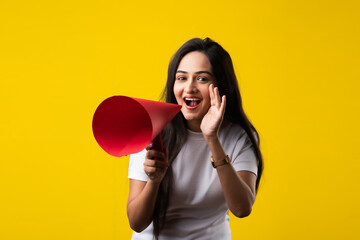 Indian pretty young woman screaming, announcing using red paper megaphone against yellow background