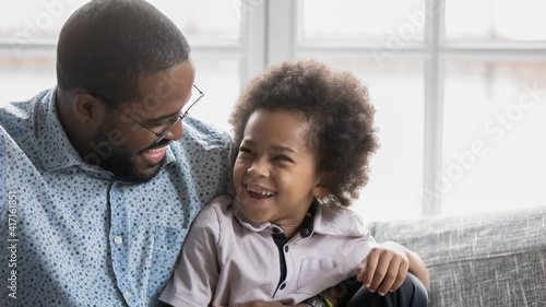 Happy cute Black boy enjoying leisure time with dad at home. Daddy hugging and tickling excited laughing little son, playing with kid on sofa in living room. Weekend with family concept. Close up
