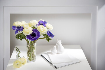 Wall Mural - Bouquet of white ranunculus and blue anemone in the vase on a white coffee table next to the fake fireplace. Shadow.