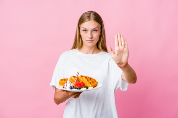 Young russian woman eating a waffle isolated standing with outstretched hand showing stop sign, preventing you.