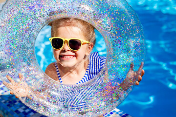 a happy little girl in sunglasses holds a swimming circle near the pool in the summer. space for text. children's summer holidays