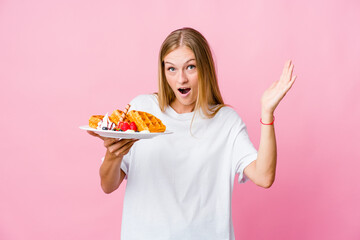 Wall Mural - Young russian woman eating a waffle isolated receiving a pleasant surprise, excited and raising hands.