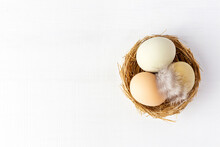 Organic Eggs In The Nest And Feather On White Wood Background, Top View, Copy Space. Three Natural Eggs In Grass Handmade Nest.