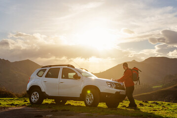 Wall Mural - Hiker with backpack looking at a map on the hood of his off-road car at sunset. Traveler and adventurous person traces his mountain route on a map. suv vehicle.