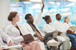 © Seventyfour - Multi-ethnic group of people wearing lab coats sitting in row in audience at medical seminar, focus on African American man raising hand to ask question, copy space