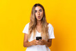 © luismolinero - Young hispanic woman over isolated yellow background looking at the camera while using the mobile with surprised expression