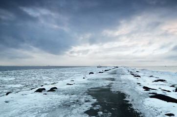Wall Mural - Empty promenade and snow-covered breakwaters close-up, frozen Baltic sea in the background. Dark storm clouds. Winter, seasons, climate change, global warming concepts. Long exposure