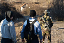Group Of Two Migrants Held By Soldier At Border Patrol On The Road In Nature In Sunny Day - Back View Of Two Unknown Men Facing Armed Man In Uniform On Road After Being Arrested For Illegal Activity