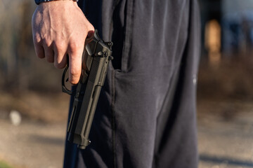 Close up on hand of unknown caucasian man holding a gun in by his leg while standing outdoor in sunny day - handgun criminal or self-defense concept with copy space