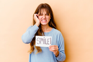 Wall Mural - Young caucasian woman holding a Smile placard isolated covering ears with hands.