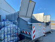 Grauer Container, ein Flügel weit geöffnet für plalttgepresste Pfandplastikflaschen in Plastikbeutel verpackt. Betonwand und strahlend blauer Himmel drum herum.