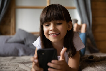 Close up of happy little Hispanic girl child lying on bed at home look at cellphone screen talk on video call. Smiling 8s Latino kid hold use smartphone play game online or study distant on gadget.