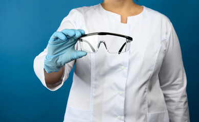 female doctor in a white medical gown stands on a blue background and holds protective plastic glasses