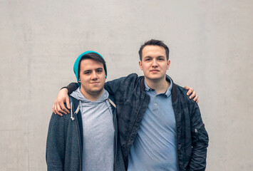 Portrait of two young people man watching the camera and embracing themselves arms on shoulders against a concrete wall. Youth lifestyle and friendship concept.