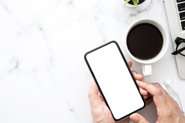 Hand holding smartphone with white blank mock up screen over white marble office desk table with laptop computer, cup of coffee and supplies. Top view image.