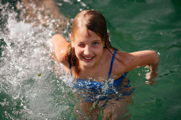 a beautiful little girl with long dark hair in a blue swimsuit swims in the outdoor pool on a hot su