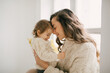 © polinaloves - Young mother hugging her little daughter, they sitting near window and smiling.