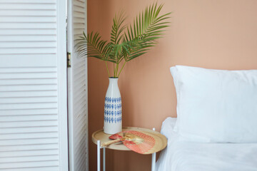 Wall Mural - Cozy interior of the white retro wardrobe, white bed and wooden table and white vase on it with green plants on the background of the peach wall