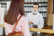 © sofiko14 - Back view of young Caucasian woman customer, paying for pills with credit card in old vintage pharmacy. Smiling senior man pharmacist swiping the credit card through payment terminal.