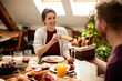 © Drazen - Happy woman talking to her husband while eating breakfast together at dining table.
