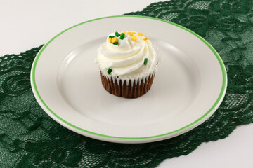 Wall Mural - Saint Patrick's Day brownie cupcake with white icing and sprinkles on white dessert late on green lace able runner on white table