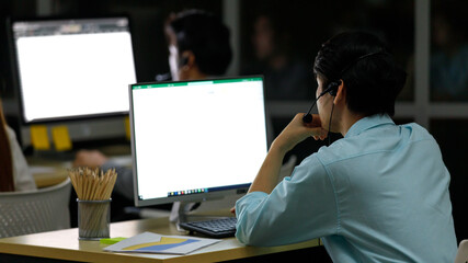 Behind Asian adult man is help desk call center do work at night to support customer and wearing headphone working at seat to look at monitor and have paper graph chart and pencils on desk