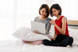 © Bangkok Click Studio - Couple adults pretty lesbians asian women hold and looking credit card and smiling feeling happy sitting on bed with white laptop on pillow on her leg after order online shopping in room on morning