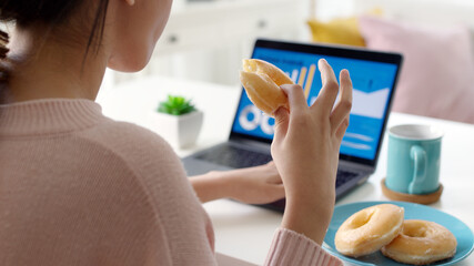 Young attractive beautiful asian female hungry eat doughnut take away snack food with full mouth look at computer notebook at home in busy work from home multitask unhealthy meal lifestyle concept.