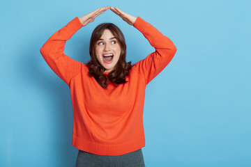 Portrait of happy excited girl wearing orange sweater feeling safe, confident under house roof gesture, dreaming of home, looking aside with opened mouth, posing isolated over blue background.