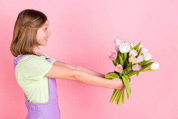 Poster - Side profile photo portrait of little girl smiling giving flowers on 8 march looking copyspace isolated on pastel pink color background