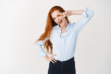 Wall Mural - Image of young ginger girl in blue shirt showing peace sign isolated over white background