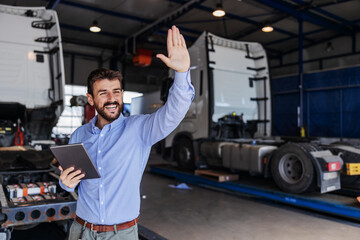 Smiling bearded chief standing in garage of shipping firm, holding tablet and waving. In background are trucks.